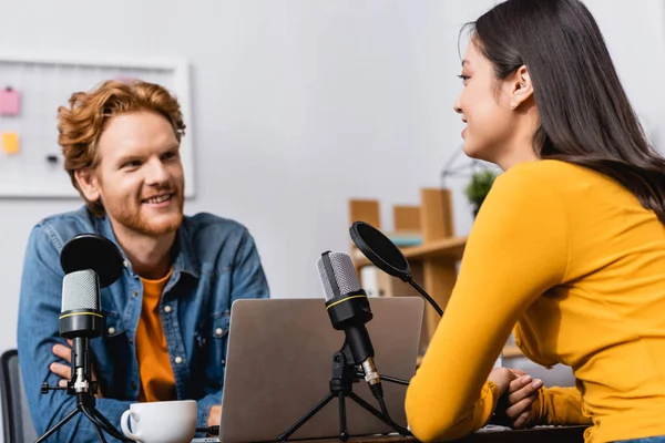 Selective focus of brunette asian woman talking to young redhead radio host during interview — Stock Photo