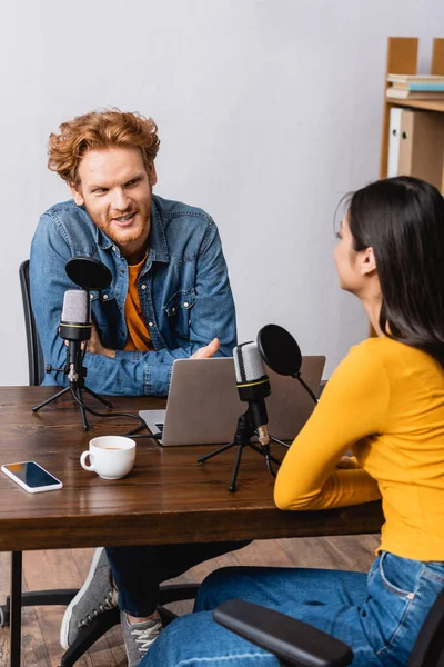 Selective focus of young redhead broadcaster talking to brunette woman during interview in radio studio — Stock Photo