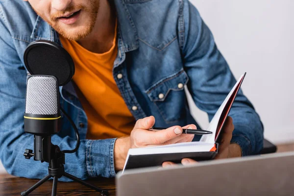 Partial view of bearded broadcaster in denim shirt holding notebook and pen while speaking in microphone — Stock Photo