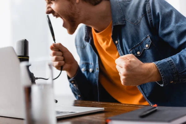 Selective focus of angry broadcaster with clenched fist screaming in microphone at workplace — Stock Photo