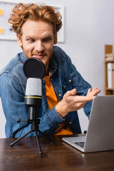 Curioso pelirrojo radio host apuntando con la mano mientras está sentado en el lugar de trabajo cerca de micrófono y portátil - foto de stock