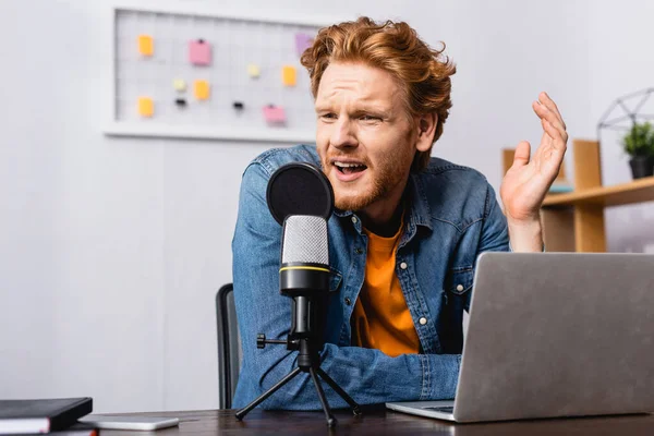 Напружений мовник в джинсовій сорочці жестикулює під час розмови в мікрофоні поблизу ноутбука — стокове фото