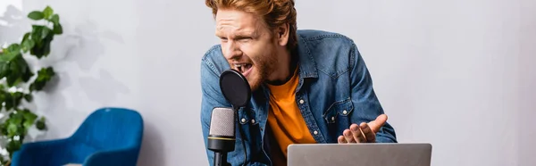 Concetto orizzontale di annunciatore rossa irritato gesticolando mentre grida nel microfono vicino al computer portatile — Foto stock