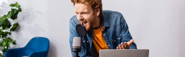 Concept horizontal de rousse irritée annonceur gestuelle tout en criant dans le microphone près de l'ordinateur portable — Photo de stock