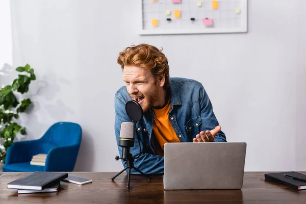 Angry redhead announcer gesturing while screaming in microphone near laptop — Stock Photo