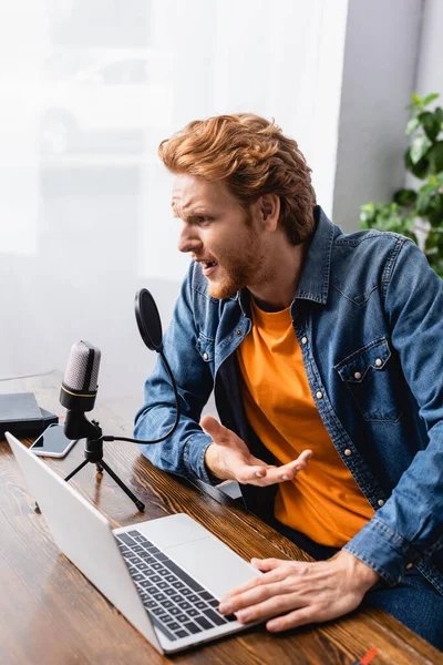 Напружений мовник в джинсовій сорочці жестикулює під час розмови в мікрофоні біля ноутбука — стокове фото