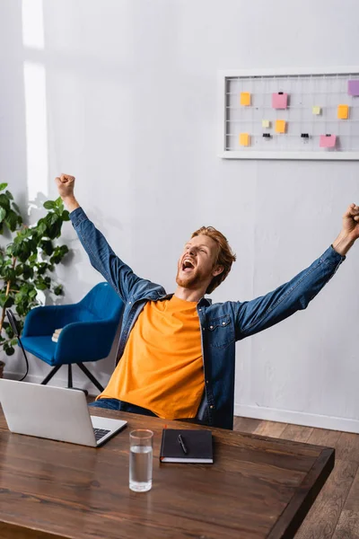 Excited radio host in denim shirt showing winner gesture and screaming near microphone and laptop — Stock Photo