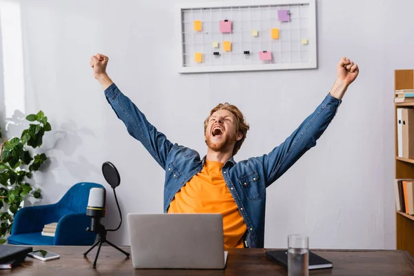 Excited radio host showing triumph gesture while screaming near microphone and laptop — Stock Photo