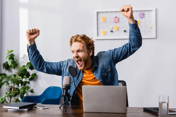 Excited announcer in denim shirt showing triumph gesture near microphone and laptop — Stock Photo