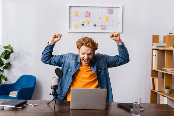 Excited broadcaster showing winner gesture while sitting at workplace near microphone and laptop — Stock Photo