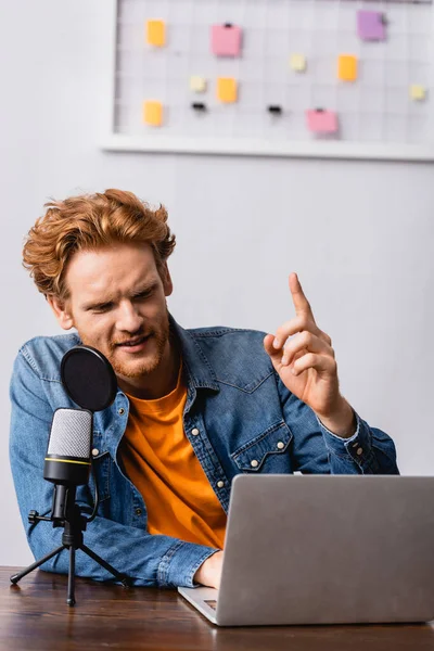 Рыжая телеведущая в джинсовой рубашке показывает жест идеи во время разговора в микрофоне рядом с ноутбуком — стоковое фото
