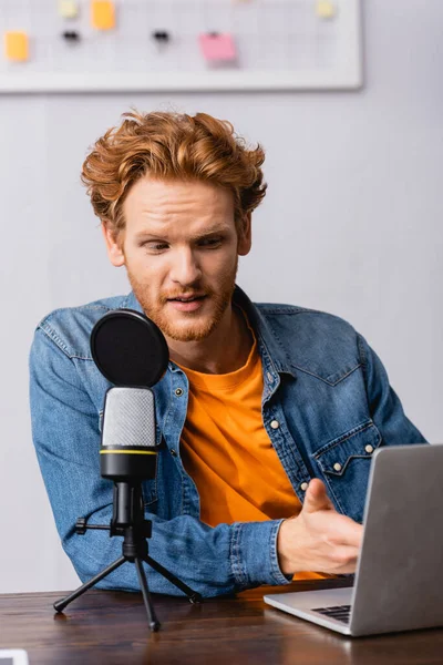 Jovem emissora em camisa jeans apontando com a mão para laptop enquanto fala em microfone — Fotografia de Stock