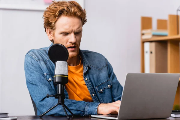 Концентрированный рыжий радиохост с помощью ноутбука, сидя рядом с микрофоном — стоковое фото