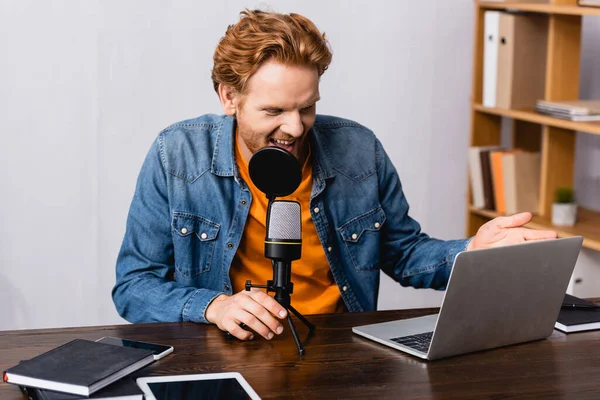 Animado, ruiva rádio anfitrião gesto enquanto fala em microfone perto de laptop e notebooks — Fotografia de Stock