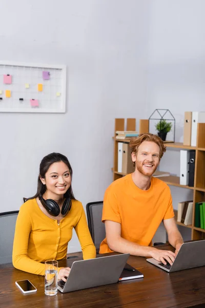 Emocionados freelancers multiculturales mirando a la cámara mientras trabajan en computadoras portátiles en casa - foto de stock