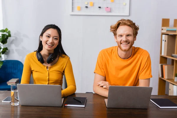 Pareja interracial de jóvenes freelancers mirando la cámara mientras están sentados cerca de computadoras portátiles y portátiles en casa - foto de stock