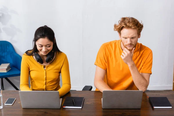 Asian freelancer with wireless headphones on neck using laptop near thoughtful man — Stock Photo