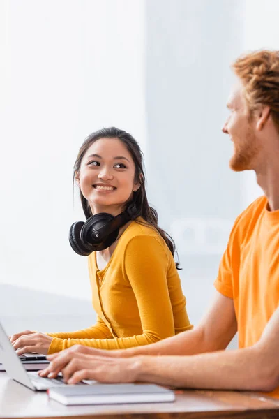Emocionado freelancer asiático con auriculares inalámbricos en el cuello mirando a un hombre joven mientras usa el ordenador portátil - foto de stock