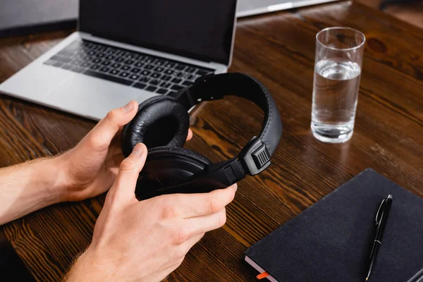Partial view of freelancer holding wireless headphones near laptop, glass of water and notebook with pen — Stock Photo