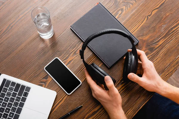 Vista cortada de freelancer segurando fones de ouvido sem fio perto de smartphone com tela em branco, laptop, notebook e vidro de água — Fotografia de Stock
