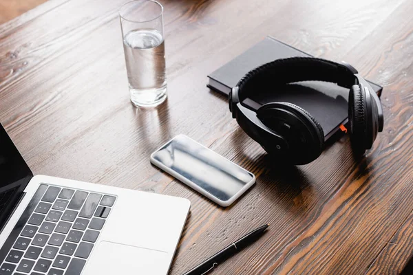Wireless headphones, smartphone, laptop, notebook and glass of water on office desk — Stock Photo