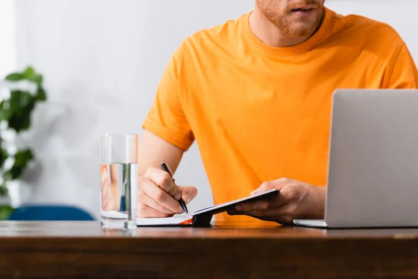 Cropped view of young freelancer writing in notebook near laptop and glass of water — Stock Photo