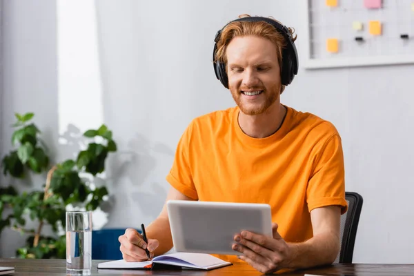 Estudante ruiva em fones de ouvido sem fio usando mesa digital enquanto escreve em notebook em casa — Fotografia de Stock