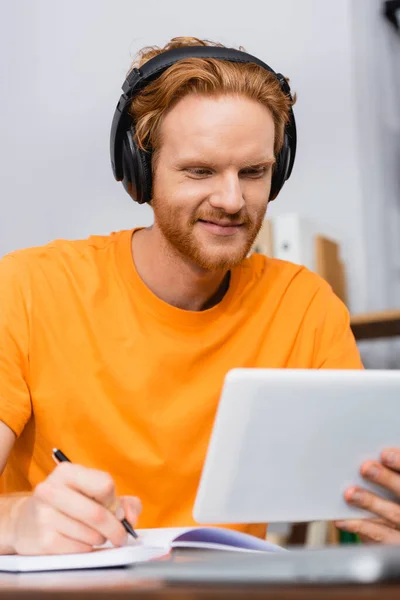 Selective focus of redhead student in wireless headphones using digital tablet and writing in notebook — Stock Photo