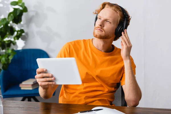 Selektiver Fokus des nachdenklichen Studenten, der drahtlose Kopfhörer berührt, während er das digitale Tablet in der Hand hält und wegschaut — Stockfoto