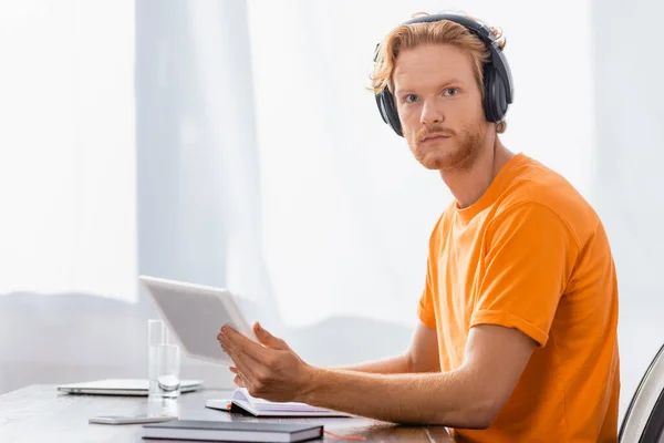 Serious student in wireless headphones holding digital tablet while looking at camera — Stock Photo
