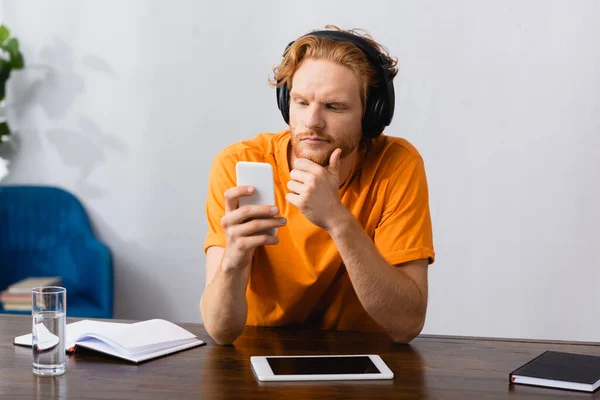 Thoughtful student in wireless headphones touching chin while using smartphone at home — Stock Photo
