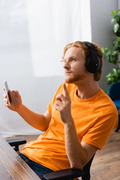 Вдумлива людина в бездротових навушниках, що показує жест ідеї, тримаючи смартфон — стокове фото