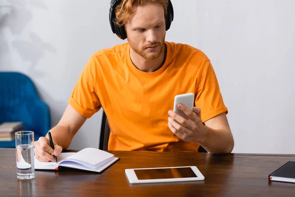 Концентрований студент в бездротових навушниках, використовуючи смартфон і пишучи в блокноті — стокове фото