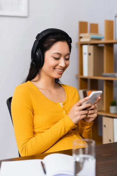 Selective focus of young asian woman in wireless headphones using smartphone near empty notebook — Stock Photo