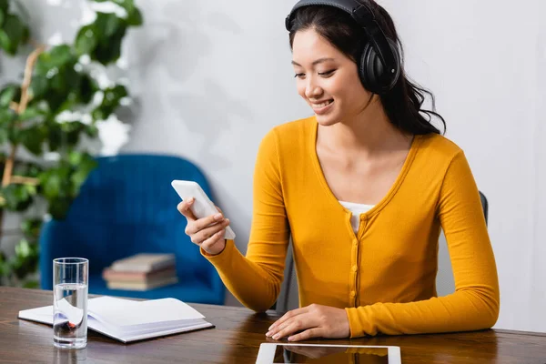 Junge asiatische Studentin in drahtlosen Kopfhörern mit Smartphone in der Nähe von digitalem Tablet, leerem Notizbuch und Glas Wasser — Stockfoto