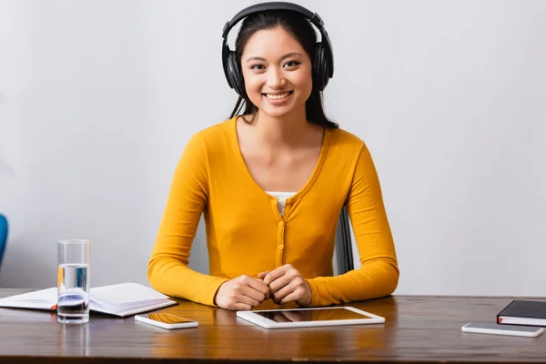 Junge asiatische Studentin mit drahtlosen Kopfhörern blickt in die Kamera, während sie am Schreibtisch in der Nähe von Geräten, Notebooks und einem Glas Wasser sitzt — Stockfoto