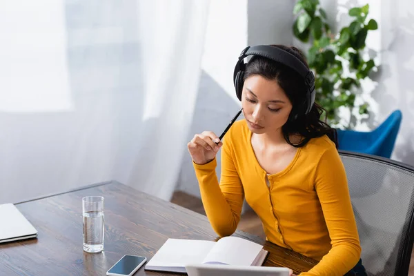 Pensativo asiático estudiante en inalámbrico auriculares celebración digital tableta y pluma cerca vacío portátil y teléfono inteligente con pantalla en blanco - foto de stock