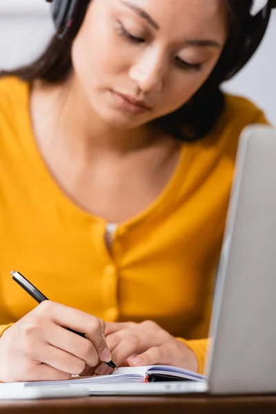 Selective focus of young asian teleworker in wireless headphones writing in notebook near laptop — Stock Photo