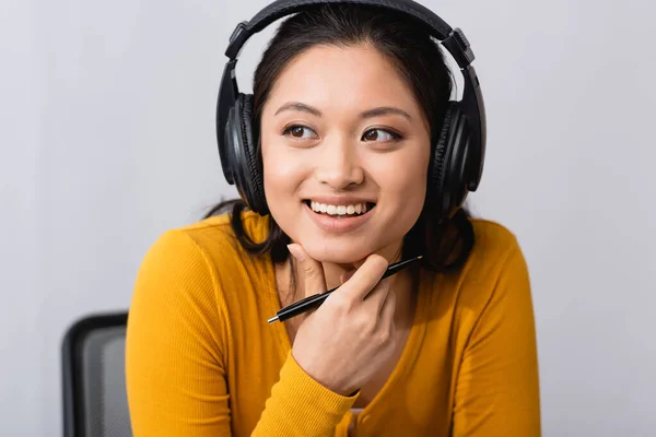 Morena mujer asiática en auriculares inalámbricos sosteniendo pluma y tocando la barbilla mientras mira hacia otro lado - foto de stock