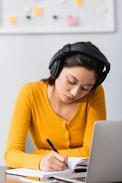 Concentrated asian teleworker in wireless headphones writing in notebook near laptop — Stock Photo