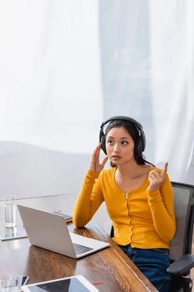 Thoughtful asian freelancer in wireless headphones showing idea sign while sitting at laptop — Stock Photo