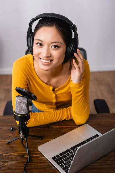 Joyeuse asiatique annonceur toucher casque sans fil tout en étant assis près du microphone et ordinateur portable en studio — Photo de stock