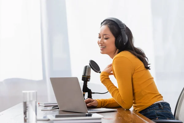 Riéndose asiático radio host en inalámbrico auriculares tocando pecho mientras trabajo cerca de micrófono y portátil - foto de stock