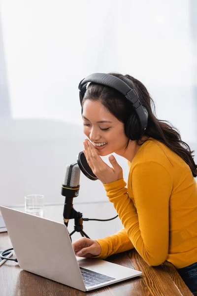 Excited asian broadcaster in wireless headphones covering mouth with hand while laughing near microphone — Stock Photo