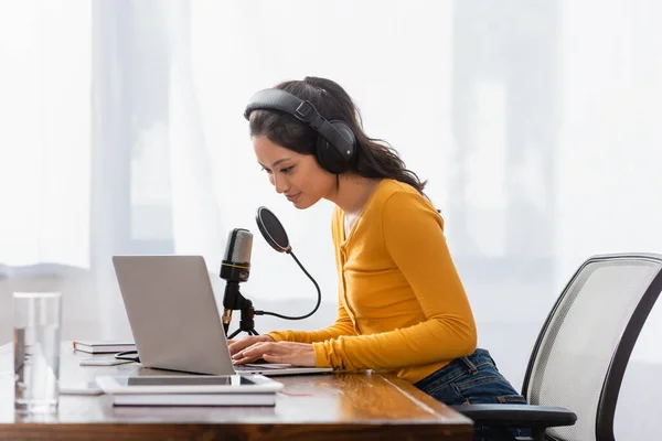 Brunette asian announcer in wireless headphones typing on laptop near microphone in studio — Stock Photo