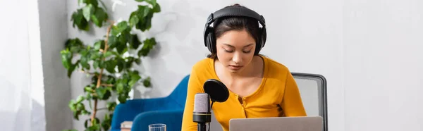 Horizontales Bild eines jungen asiatischen Radiomoderators mit drahtlosen Kopfhörern, der in der Nähe von Mikrofon und Laptop im Studio arbeitet — Stockfoto