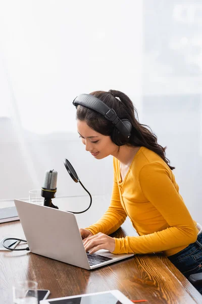 Foyer sélectif de l'annonceur asiatique dans les écouteurs sans fil tapant sur ordinateur portable près du microphone — Photo de stock