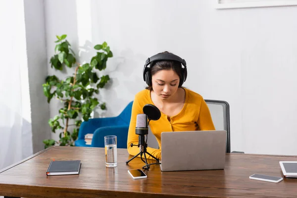 Brunette asian broadcaster using laptop near microphone, glass of water and gadgets at workplace — Stock Photo