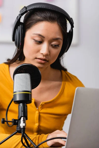 Selective focus of concentrated asian broadcaster in wireless headphones using laptop near microphone — Stock Photo