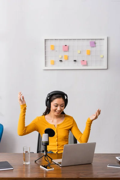 Excité diffuseur asiatique dans les écouteurs sans fil montrant wow geste tout en travaillant près de microphone et ordinateur portable — Photo de stock